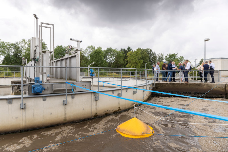 Foto (Außenaufnahme) eiens Teils der biologischen Reinigungsstufe einer Modell-Kläranlage. Ein Steg führt über das Reinigungsbecken, auf dem Steg steht eine Gruppe von Personen die an einer Fachexkursion teilnehmen. Das Becken durchfließt (braunes) Abwasser. An der Abwasseroberfläche treibt eine gelbe Messhaube die dort verschnürt ist. Die Messhaube dient der Echtzeitmessung von (unsichtbaren) Treibhausgasen die aus dem Becken austreten können.